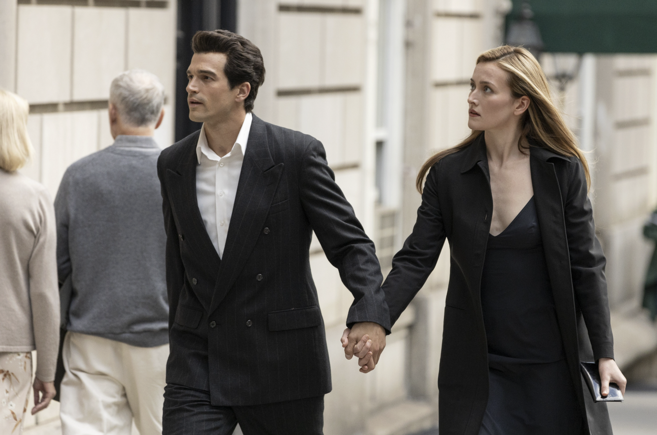 « Photographie des acteurs interprétant le couple Kennedy-Bessette dans la série "Love Story" (Disney+, février 2026). L'actrice incarnant Carolyn porte une robe minimaliste noire assortie d'une veste structurée et d'une pochette sobre, tandis que l'acteur jouant John F. Kennedy Jr. figure à ses côtés. Ce visuel symbolise l'esthétique Calvin Klein des années 90, moteur de l'explosion des requêtes Google sur le luxe discret ce mois-ci. »