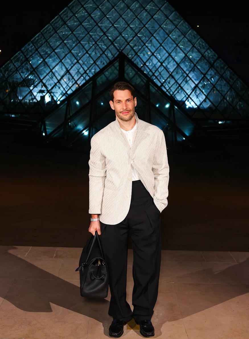 Simon Porte Jacquemus, souriant, pose devant la Pyramide du Louvre illuminée à la tombée de la nuit. Il porte un costume noir épuré et tient à la main un sac.À l'arrière-plan, la structure en verre de la Pyramide se découpant sur le ciel sombre.